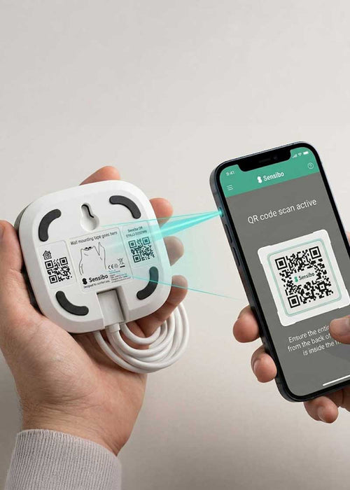 PROMPT: Hands holding phone scanning QR code on back of small white Sensibo device, blurred modern apartment background, warm natural light, step-by-step tutorial feel