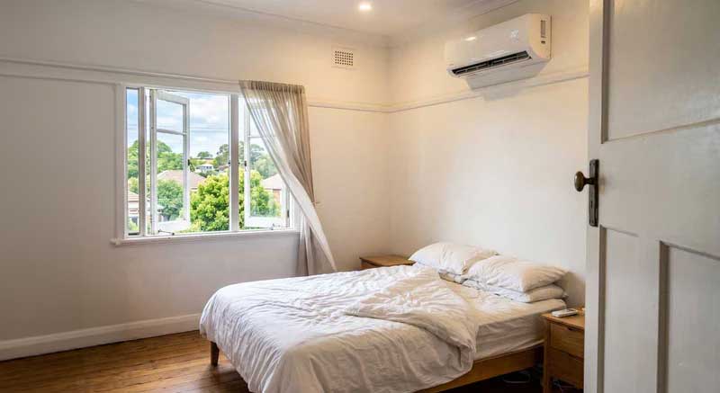 PROMPT: Split AC unit running on wall with frost/ice buildup, open balcony door with curtains blowing in, bright summer day outside, energy waste concept, editorial photography style